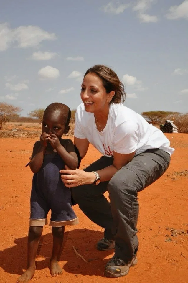 Layla Dean‑Verity, founder of Advocacy AI and faculty & contributors member, crouching beside a barefoot child on reddish‑orange soil in a semi‑arid landscape under a partly cloudy sky. Professional humanitarian‑style portrait symbolising leadership, compassion, and advocacy.