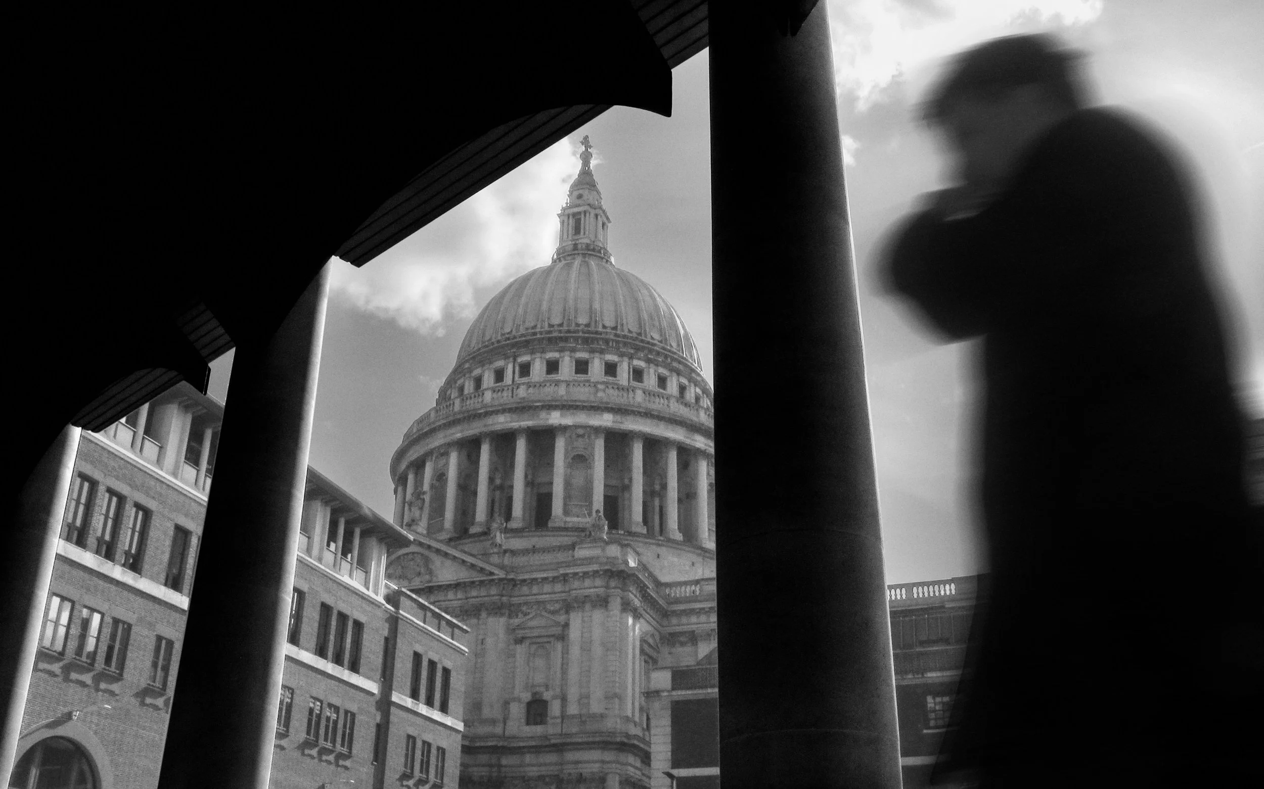 St. Paul’s Cathedral dome in London, black-and-white photograph framed by foreground columns and arches, blurred figure walking on right adding motion contrast, dramatic composition highlighting classical architecture and iconic landmark.