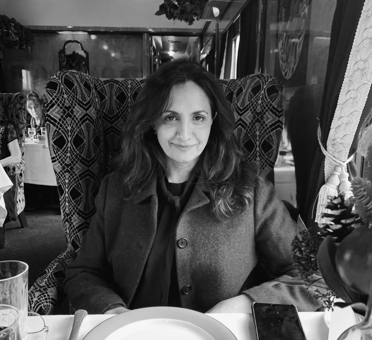 Meet the founder of Advocacy AI, Layla Dean-Verity, seated at a dining table in a vintage-style train carriage or restaurant, wearing a dark coat with long hair, black-and-white photograph with patterned high-back seating chairs, lace curtains, floral arrangements, and elegant dining setting, timeless professional portrait style.
