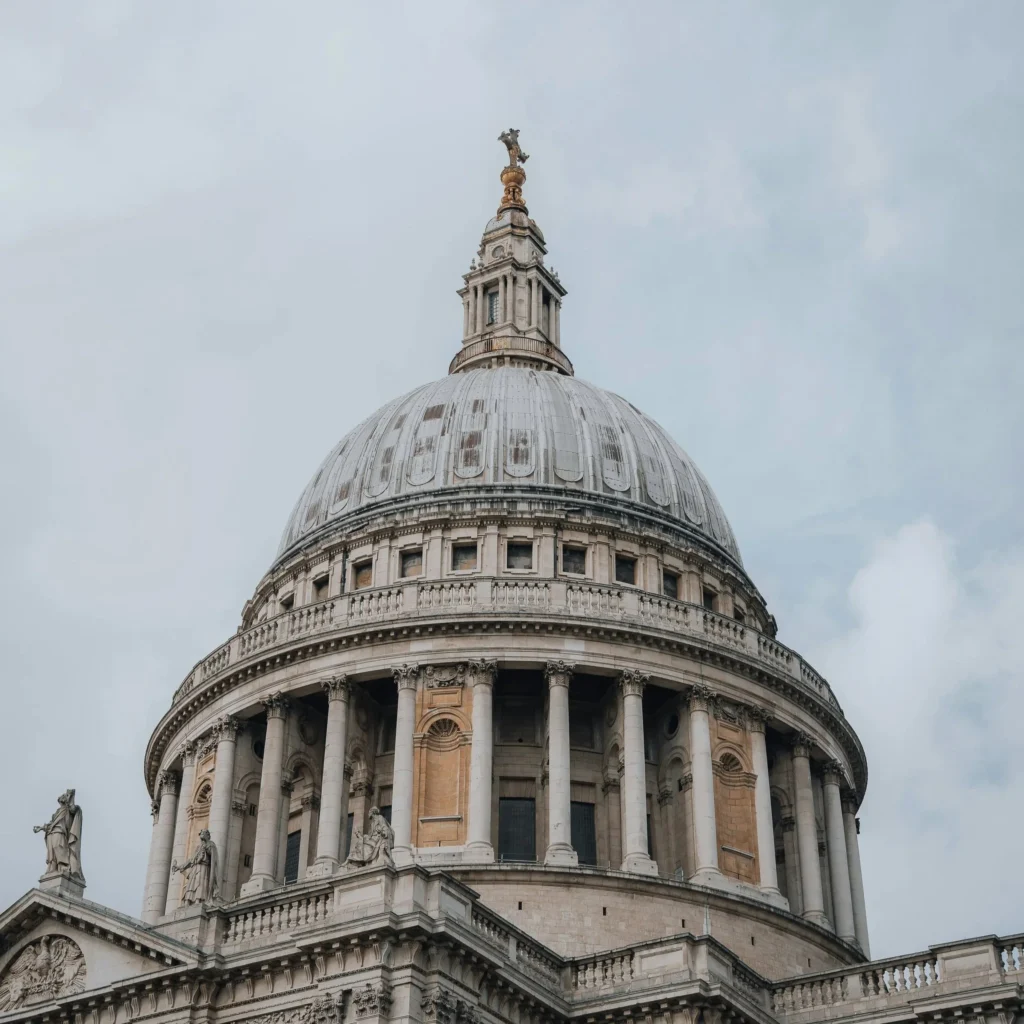 St. Paul’s Cathedral dome in London, English Baroque architecture with large stone dome topped by lantern and golden statue, rows of Corinthian columns and ornate carvings below, symmetrical classical design in light-colored stone, iconic historical landmark.