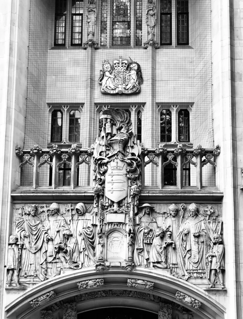Supreme Court of the United Kingdom façade in London, Gothic Revival architecture with intricate stone carvings and sculptures. Royal Coat of Arms featuring lion and unicorn flanking crowned shield at the top, detailed reliefs of historical and allegorical figures below, and central heraldic shield with three swords. Elaborate decorative elements symbolise law, authority, and tradition.