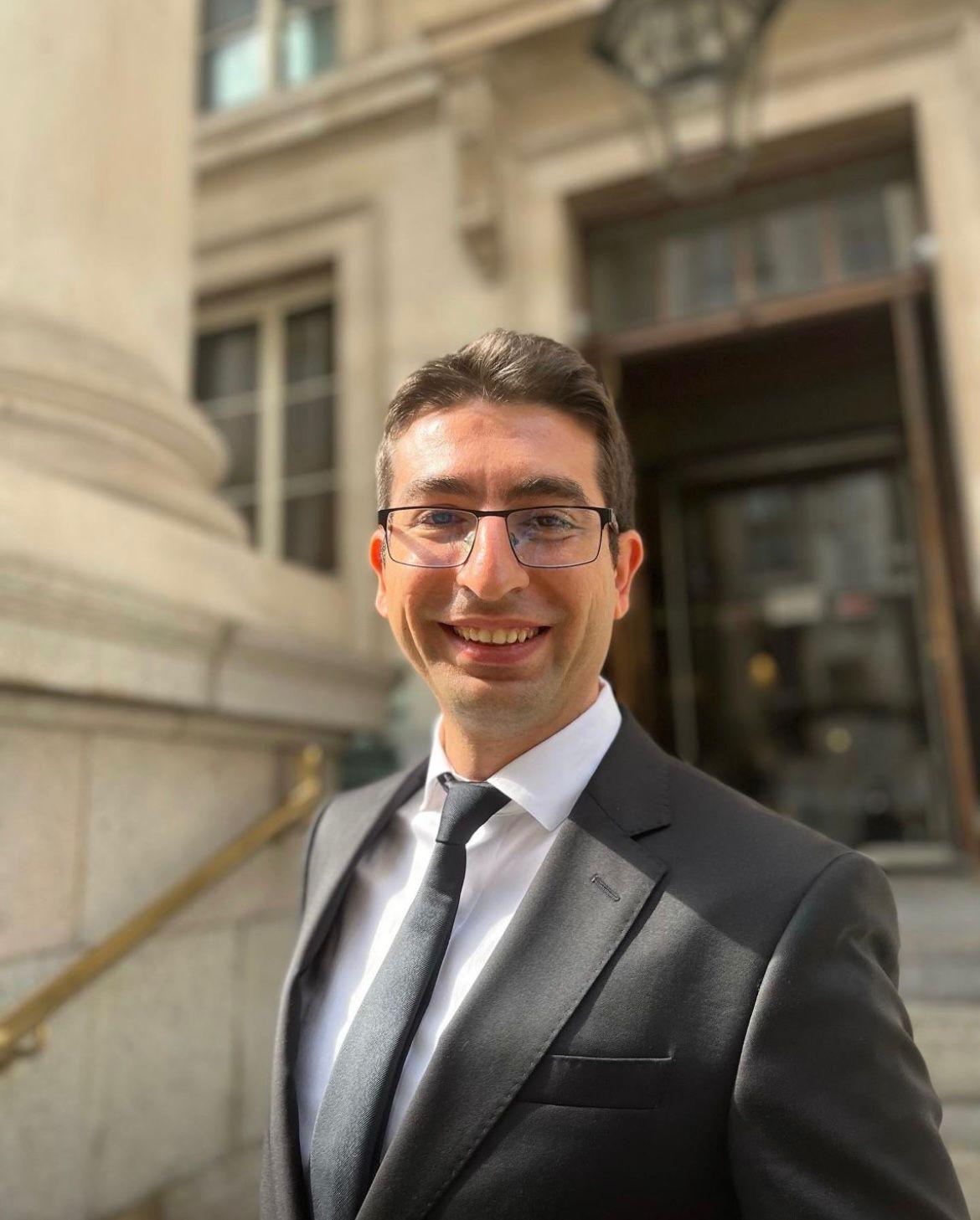 Can Eken, Advocacy AI team member, in a dark suit with white shirt and dark tie, wearing glasses, standing outdoors in front of a stone building with classical columns, lantern, and brass handrail, formal professional portrait style.