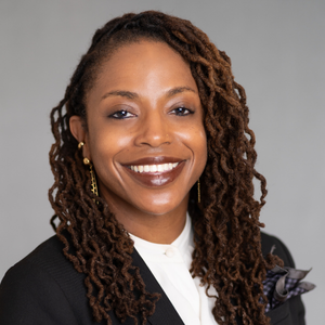 Diona Howard-Nicolas, Advocacy AI team member, in a dark suit jacket with white collared shirt and patterned pocket square, long styled dreadlocks, against a plain gray background, formal portrait style.