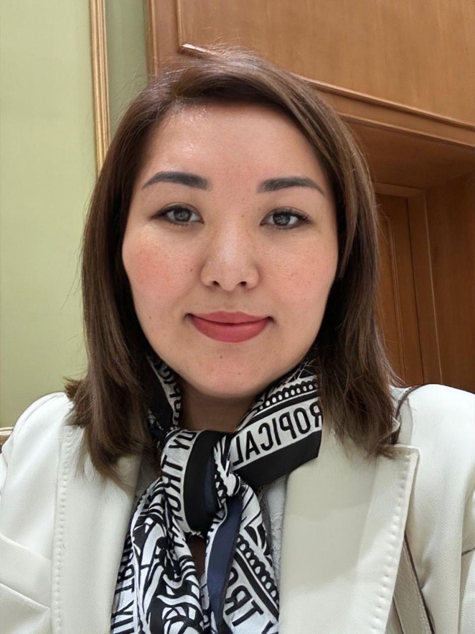 Meet The Team 9 Assel Upetova, Advocacy AI team member, wearing light blazer and patterned black‑and‑white text scarf. Shoulder‑length brown hair, standing indoors against wooden panel and light green wall, professional portrait emphasising professionalism and approachable style.