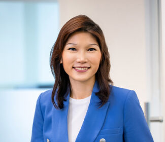 Charis Tan, Advocacy AI team member, in a bright blue blazer over a white top, standing in a professional office setting with light walls and glass partition, formal portrait style.