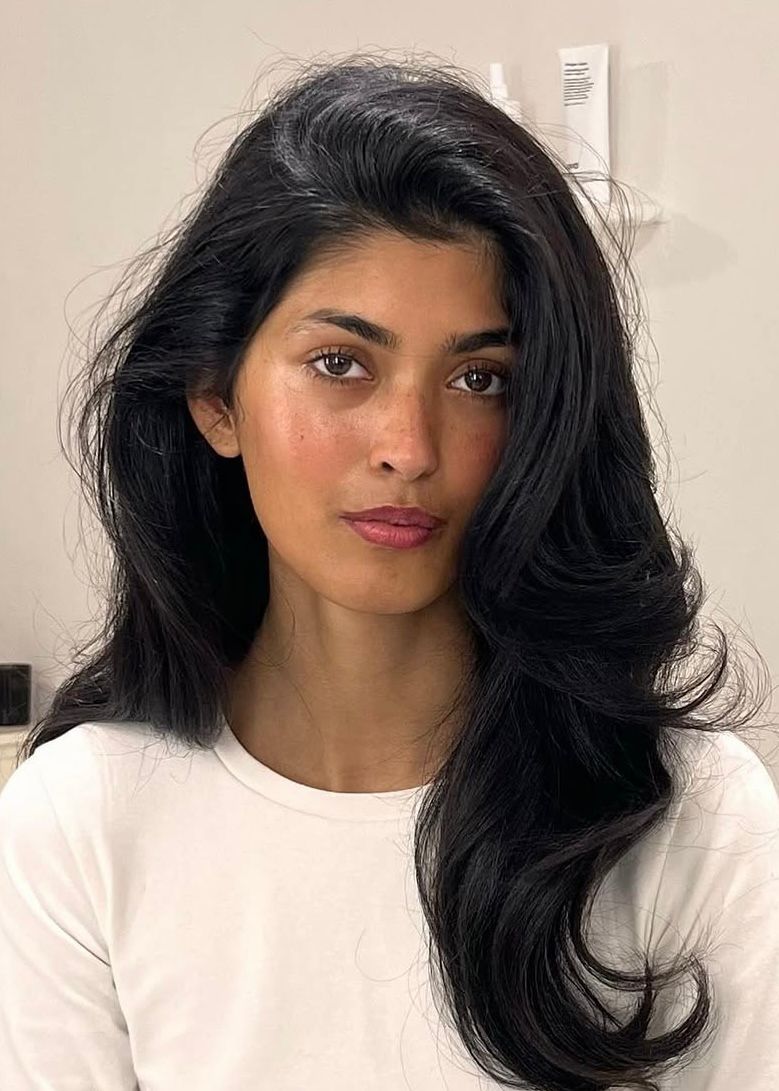 Meet The Team 14 Naina Hamayoun portrait with long wavy black hair, wearing white top. Calm expression while looking directly at camera, set against neutral indoor background. Professional image emphasising poise, approachability, and team presence.