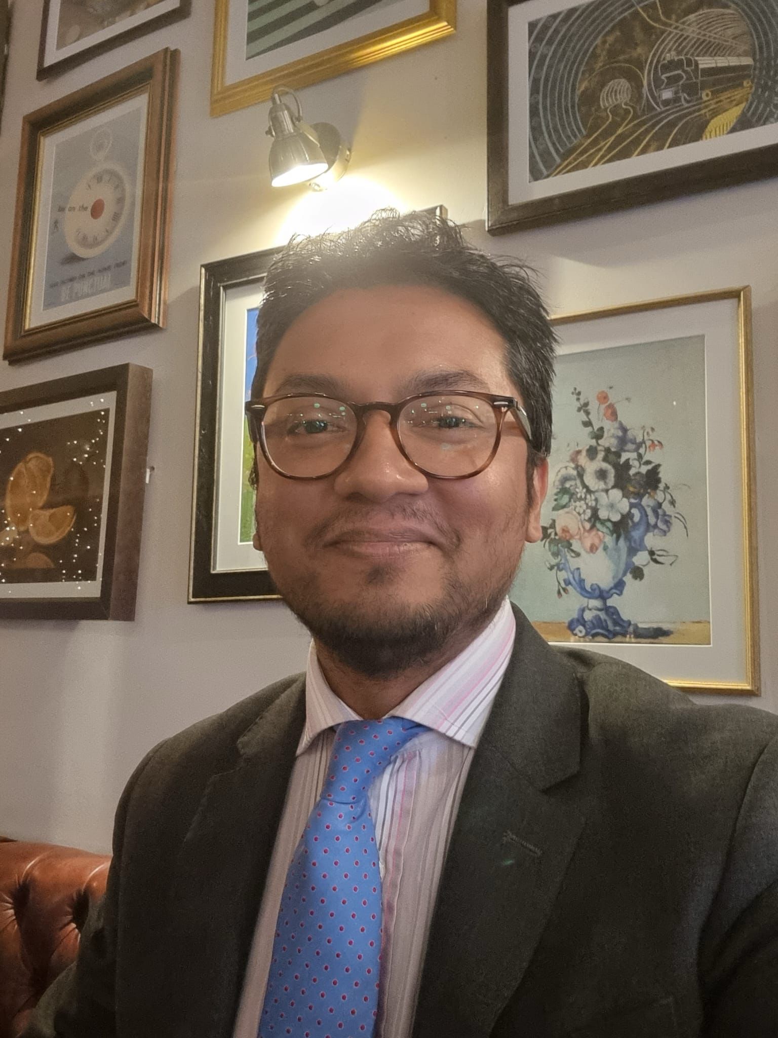 Meet The Team 5 Reza Choudhury, Advocacy AI team member, in dark suit jacket, light pink shirt, and blue tie with pink dot pattern. Seated on brown leather couch indoors with framed artworks and wall‑mounted light fixture, professional portrait emphasising expertise and approachable style.