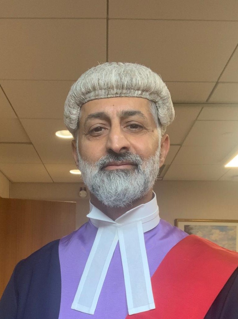 Meet The Team 4 Tahir Khan, Advocacy AI team member, wearing formal judicial attire including white wig, purple robe, red sash, and white neck band. Professional portrait indoors with recessed ceiling lights and framed wall art, emphasising authority, tradition, and legal expertise.